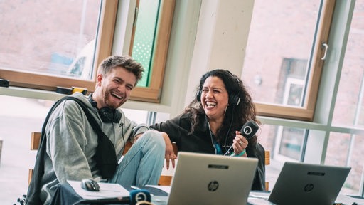 Zwei Mitarbeitende von Radio Bremen sitzen vor Laptops und lachen.