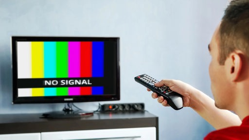 Ein Mann sitzt mit einer Fernbedienung vor dem Fernseher, auf dem "No Signal" steht (Symbolbild)