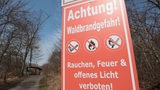 Auf einem Schild steht: "Achtung! Waldbrandgefahr!" Im Hintergrund ist ein Waldstück zu sehen.